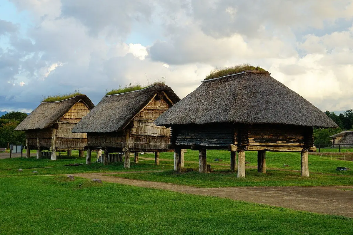 Three reconstructed raised-floor buildings with thatched roofs standing on a lawn.