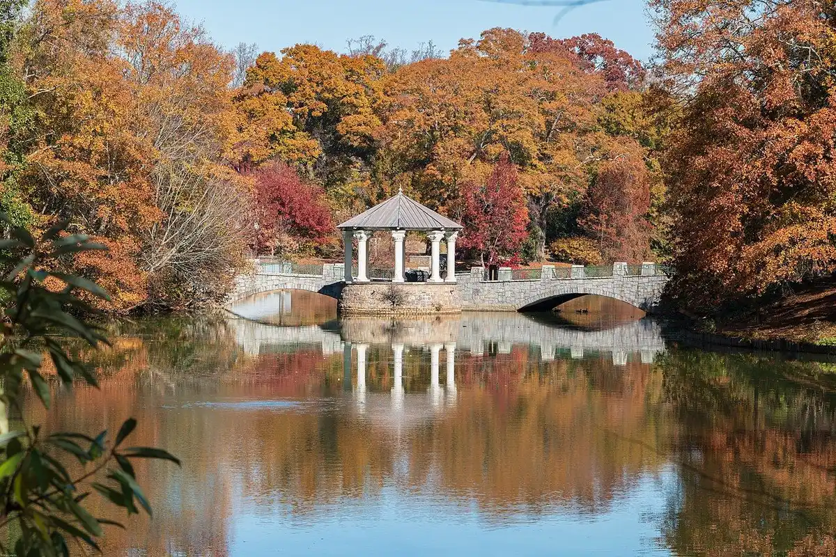 A white gazebo and stone bridge on the lake in Piedmont Park, surrounded by autumn trees and reflected in the water.