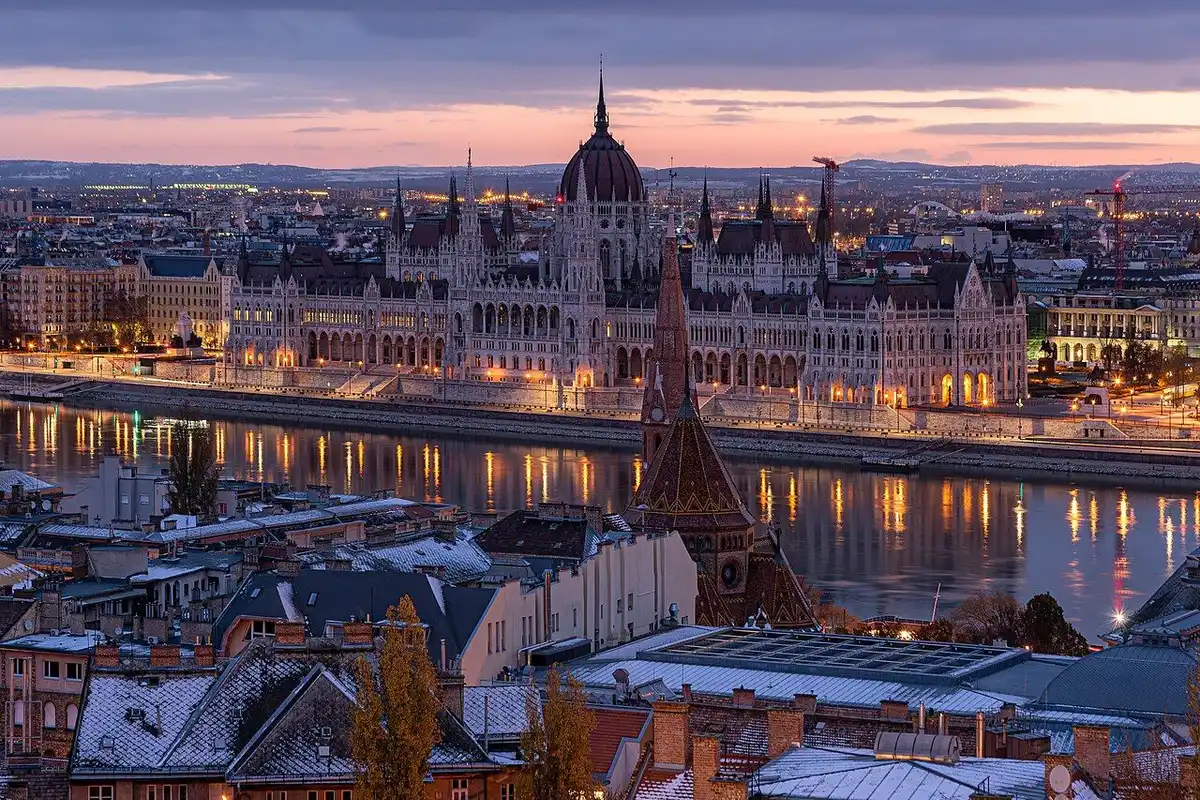 The lit Parliament reflected in the Danube at dawn, beyond snow-covered rooftops on the Buda side.