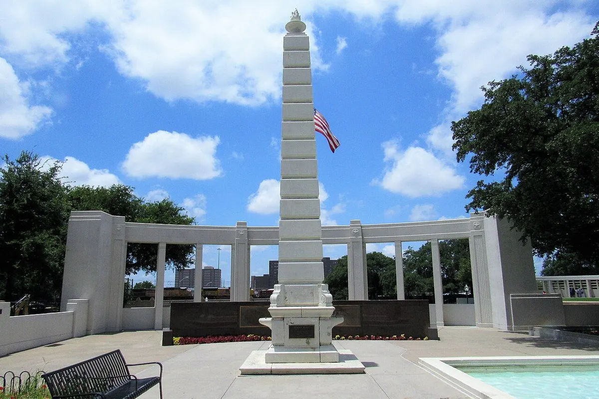 The white memorial obelisk and curved colonnade at Dealey Plaza in Dallas, with the U.S. flag and a bench.