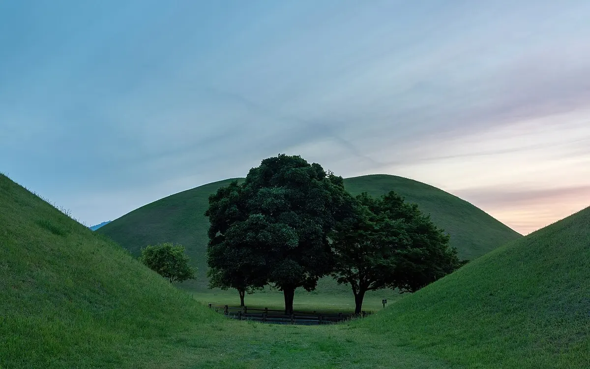 Grass-covered Silla tumuli and trees under a colourful sunset sky at Daereungwon Tomb Complex in Gyeongju.
