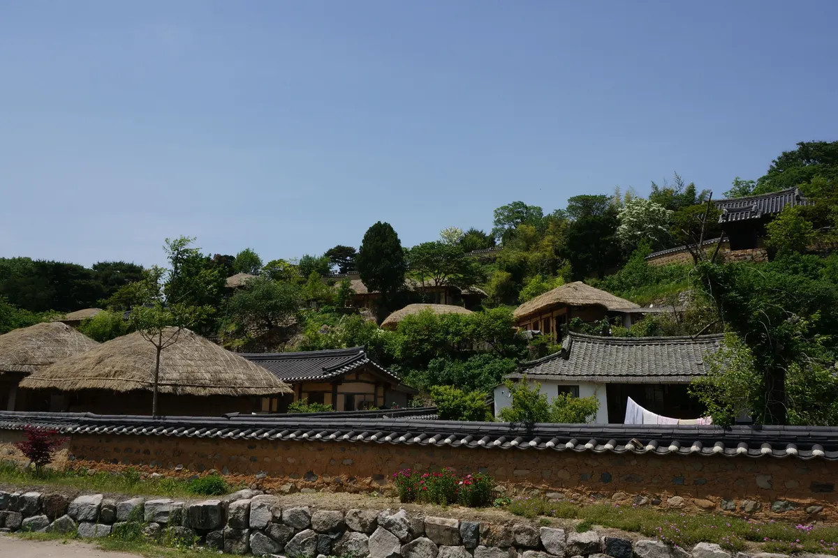 Beyond a tile-capped stone wall in Yangdong Village, thatched and tiled houses are scattered across a green hillside.
