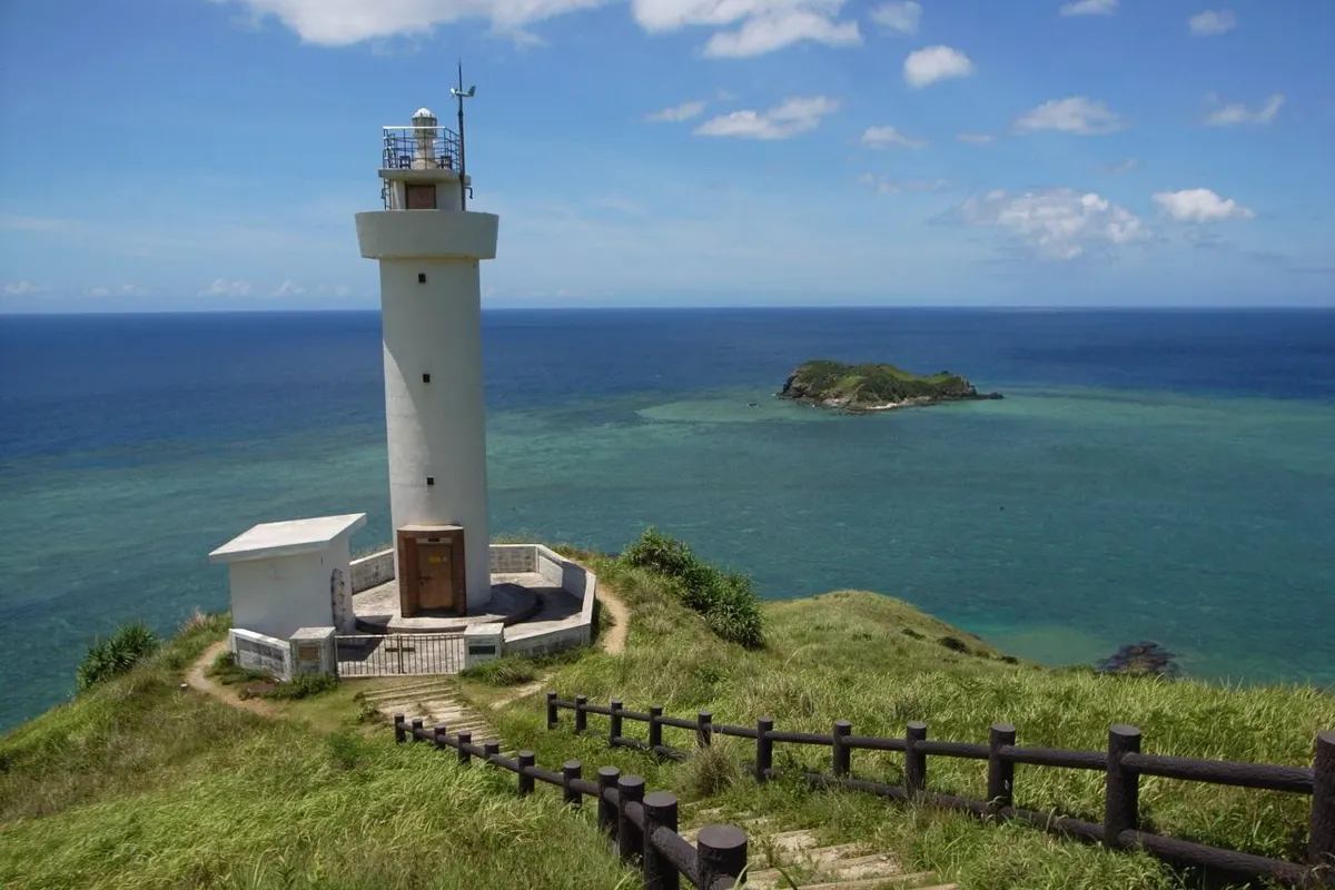 The white Hirakubo-zaki lighthouse on a grassy headland above a turquoise coral sea, with a small islet offshore.