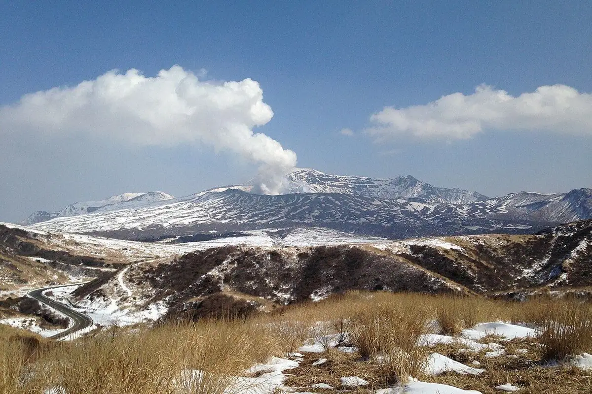 White steam rises into a blue sky from snow-covered Mount Aso Nakadake, with snow lingering on dry grass in front.