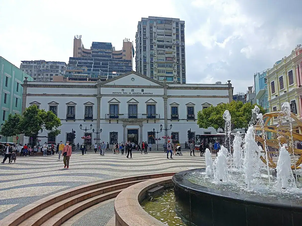 Wave-patterned mosaic pavement and a fountain in front of a white neoclassical municipal building