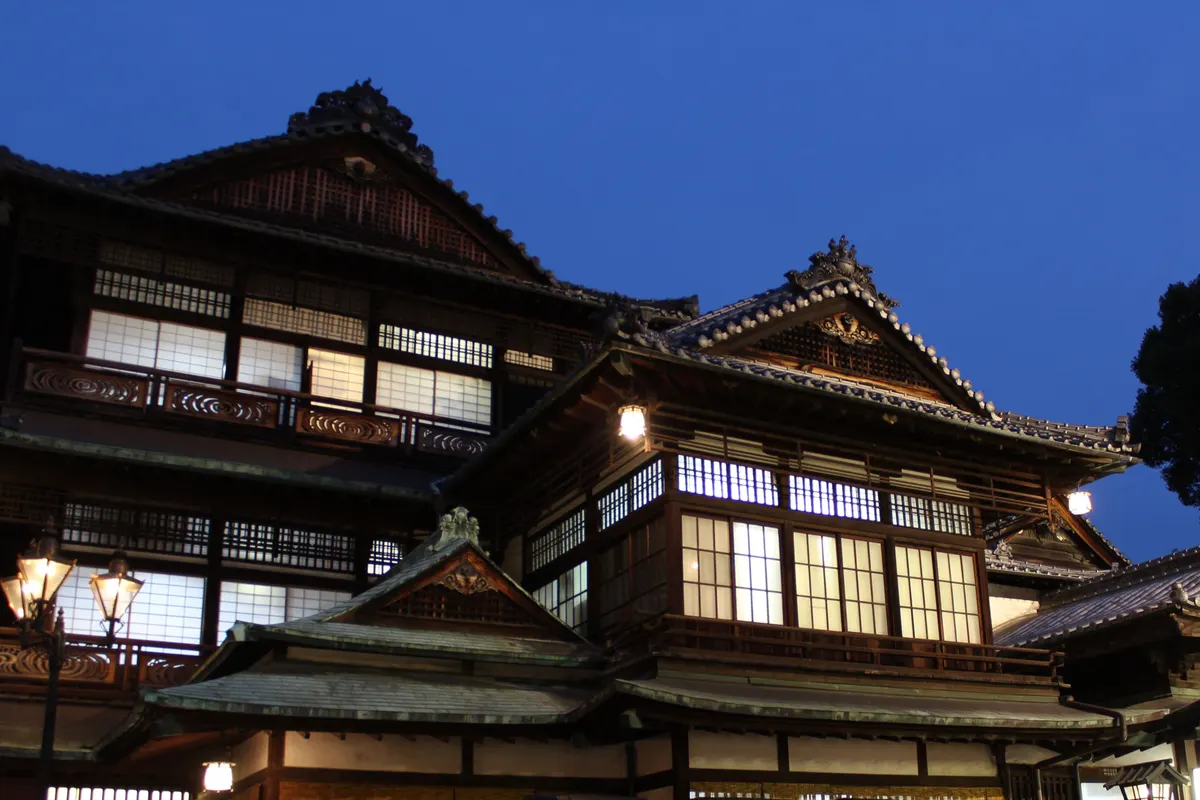 Warm light glows from the windows of the wooden Dogo Onsen Honkan under a deep blue evening sky.