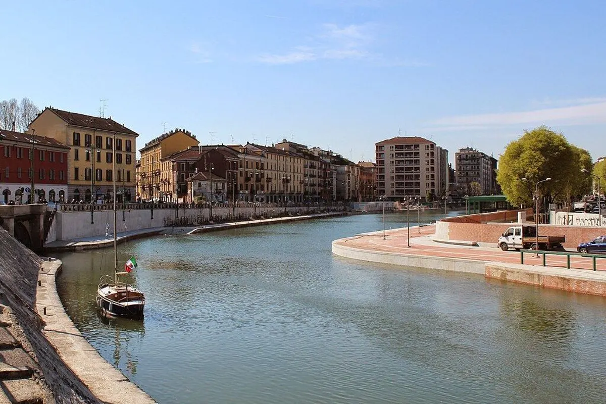 A small sailboat on the calm water of the Darsena under a blue sky, with old buildings along the far bank.