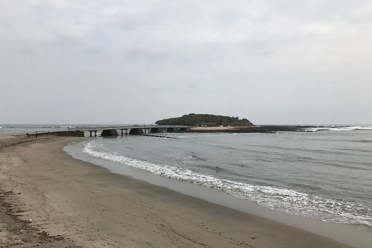 The small wooded island of Aoshima, linked by a bridge, seen across a sandy beach.