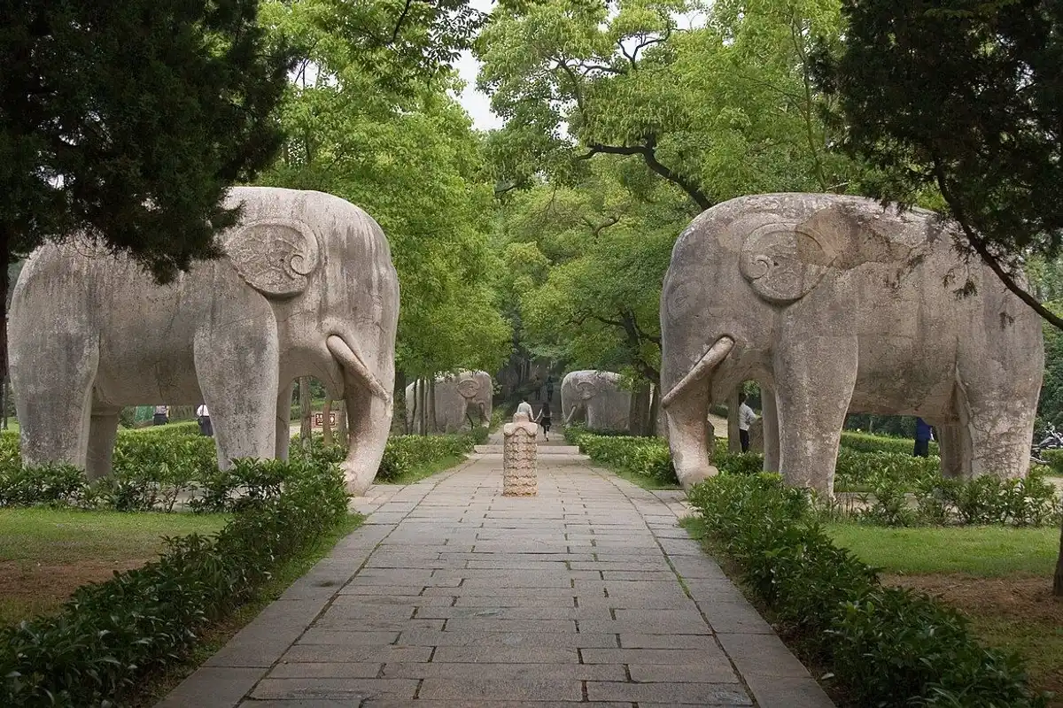 Large stone elephants facing each other on both sides of a tree-shaded stone path.