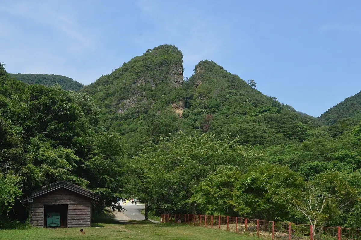 Doyu no Warito, the V-shaped split summit of the Sado Gold Mine, rising above green forest, with a small wooden hut on a lawn in front.
