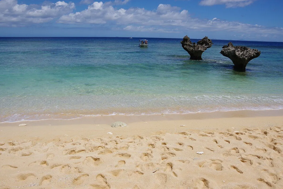 Two heart-shaped rocks standing in clear emerald water off a sandy beach.