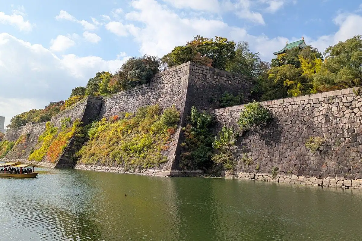 A tour boat passing along the green moat below huge stone walls, with the main tower's roof above the trees.