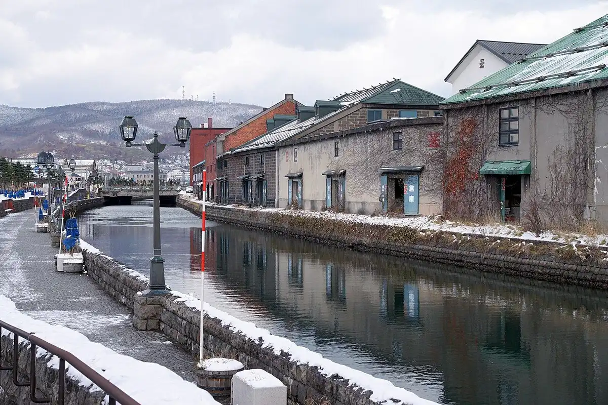Old stone warehouses and a snow-dusted promenade line the Otaru Canal beneath snowy hills.