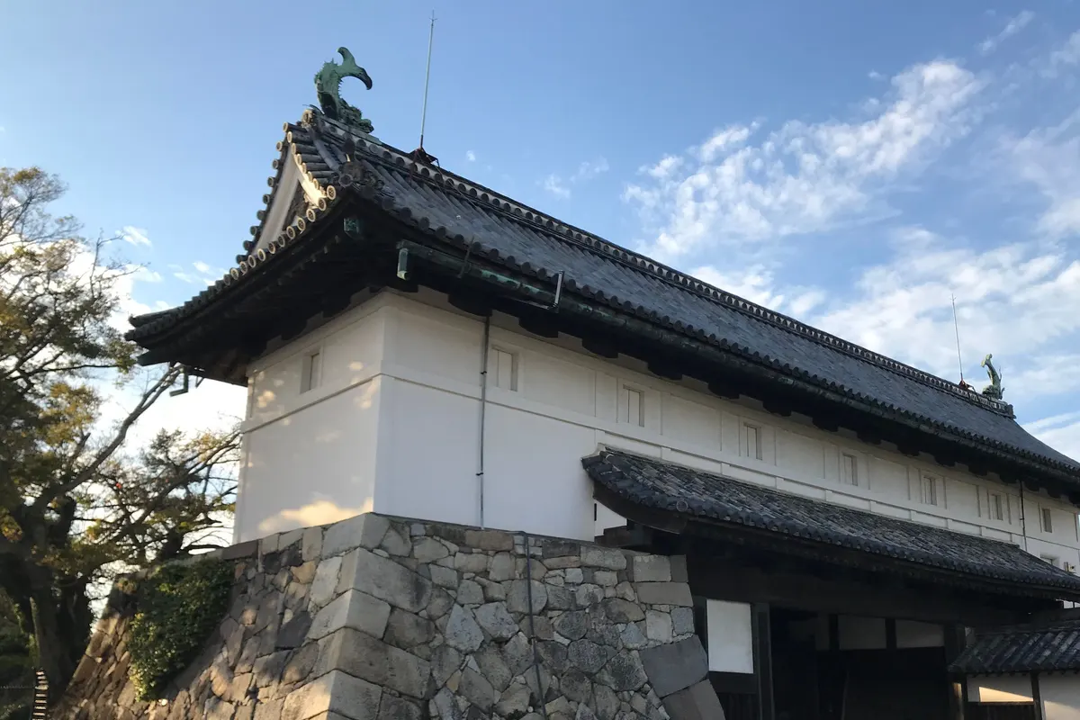 Saga Castle's Shachinomon Gate stands on a massive stone base in autumn afternoon light, with bronze shachi ornaments on both ends of its tiled roof.