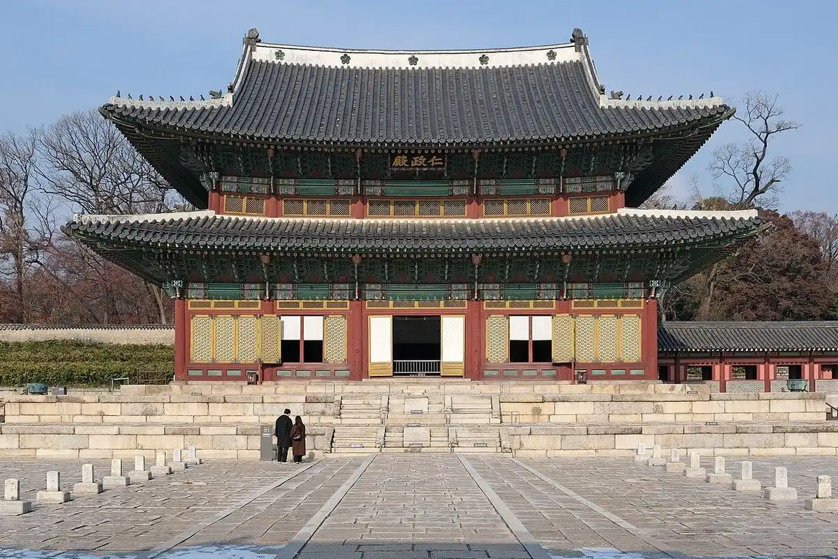 Injeongjeon, the main throne hall of Changdeokgung Palace, rises with its double roof on a raised stone terrace at the end of a wide stone courtyard, backed by late-autumn trees.