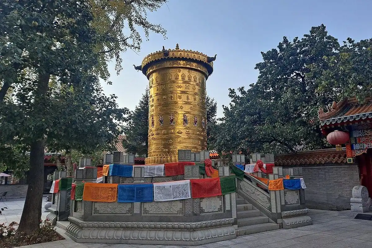 A huge golden prayer wheel on a stone base hung with colourful prayer flags, surrounded by green trees.