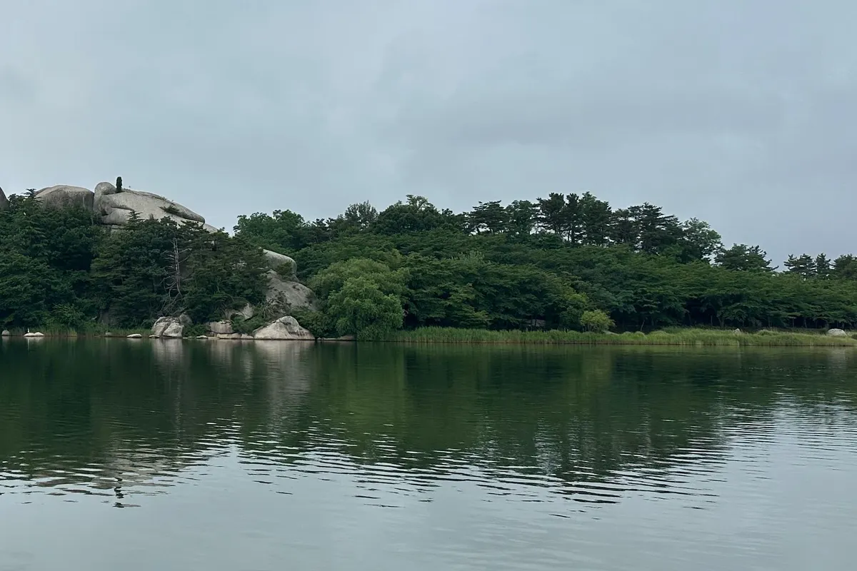 Large granite boulders and pine woods line the calm shore of Yeongnangho Lake in Sokcho, reflected in the water under a cloudy sky.