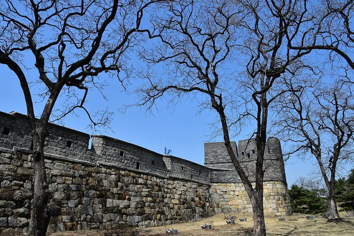 The stone walls and a rounded bastion of Hwaseong Fortress stretch between bare trees under a blue sky.