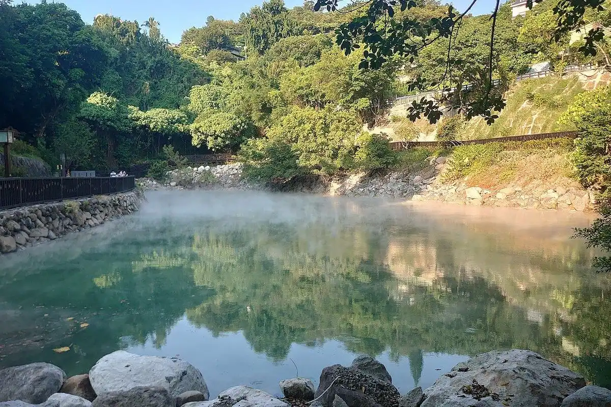 Steam rising from the jade-green hot spring pond, with the green forest reflected in the water.