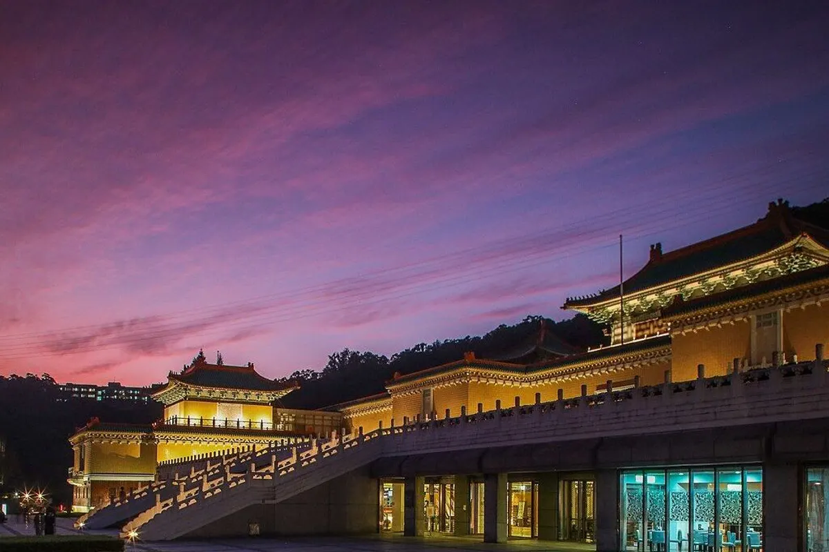 The lit halls and white balustraded stairs of the National Palace Museum under a purple sunset sky.