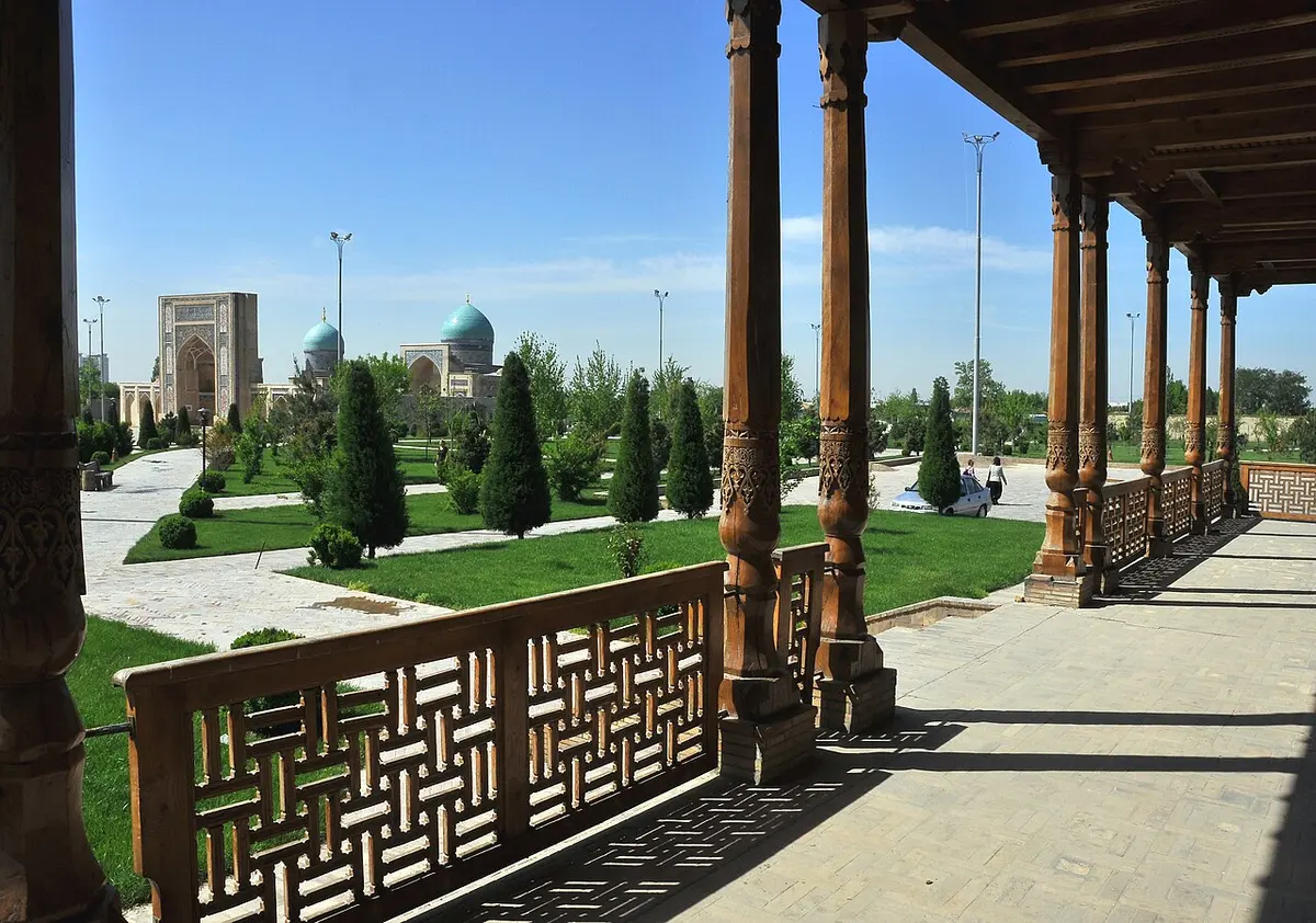 The Khazrati Imam complex in Tashkent seen from a loggia of carved wooden columns, with gardens and turquoise-domed buildings beyond.