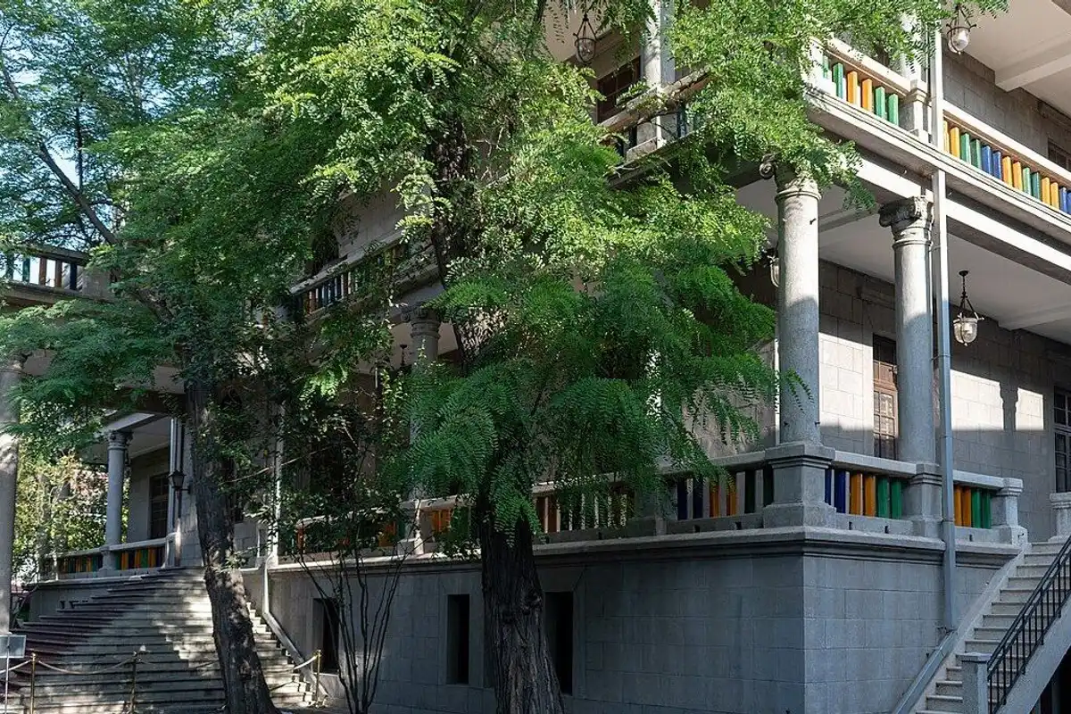 A historic villa with a grey stone colonnaded veranda and outdoor stairs, seen through green trees.