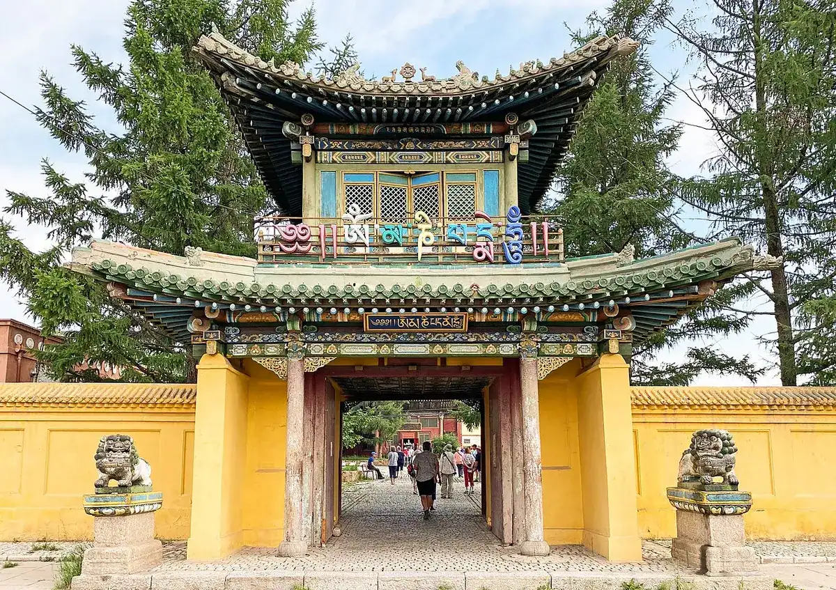 Monastery gate with a green tiled roof and yellow walls, flanked by stone lion statues, with visitors walking through toward the courtyard
