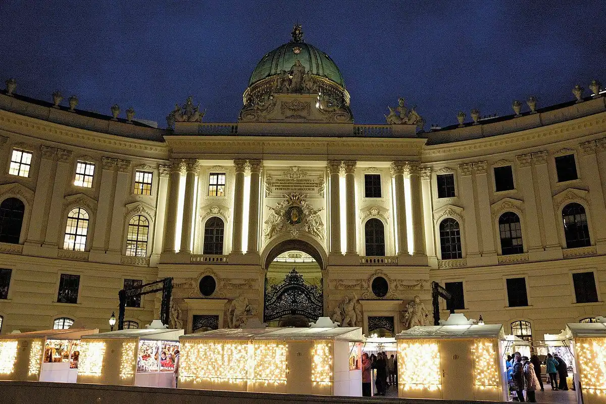 Light-strung stalls lined up in front of the illuminated Hofburg under the evening sky.