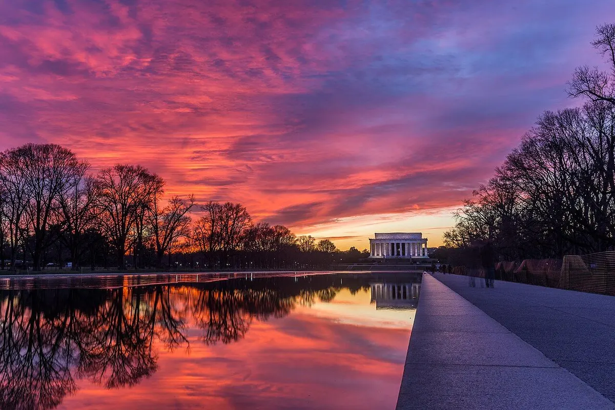 The Lincoln Memorial at the end of the Reflecting Pool, with a red sunset sky mirrored in the water and bare trees on both sides.