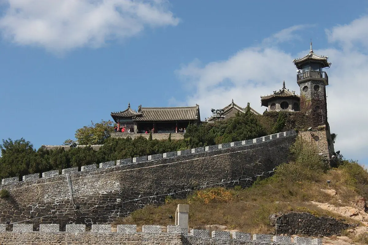 Penglai Pavilion's halls and tower along an old wall on a hilltop under a blue sky.