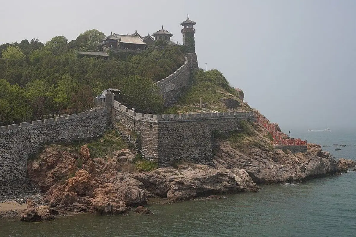 An old stone wall running up reddish sea cliffs to the halls and tower on the hilltop, with the sea alongside.