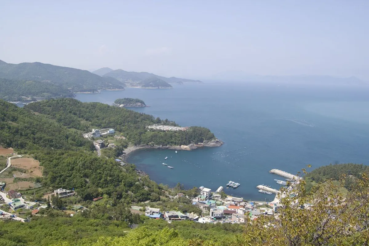 A small fishing harbour below green hills, with a blue bay and layered islands in the distance.
