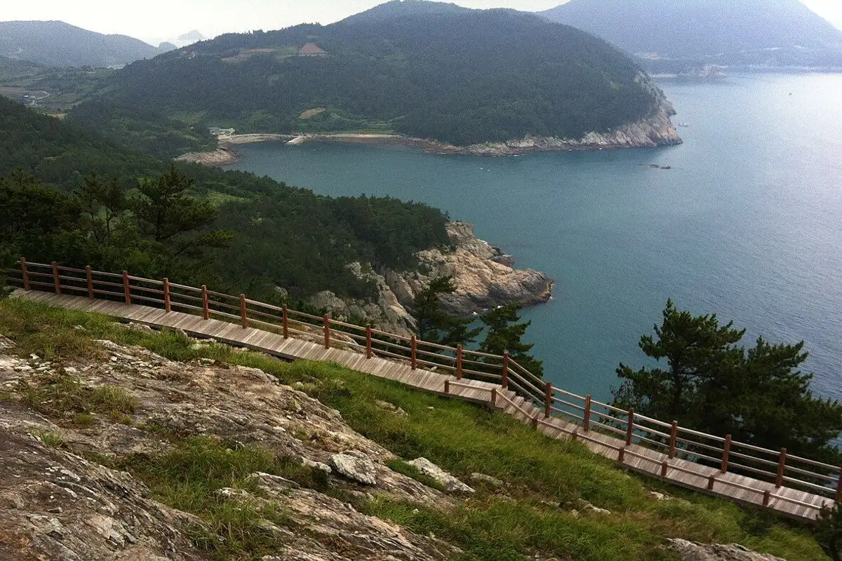 A wooden boardwalk running along sea cliffs above a blue bay and green, forested islands.