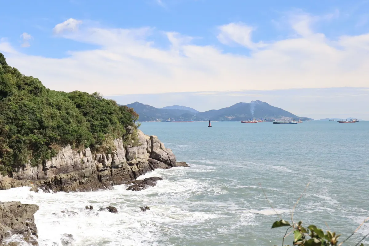 Waves break on the forested rocky shore of Odongdo Island, with islands and ships out at sea.