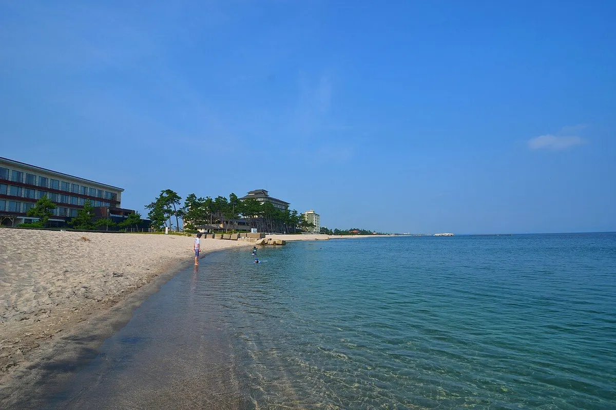 Clear water and a white-sand beach at Kaike, with pines and hot spring hotels along the shore.
