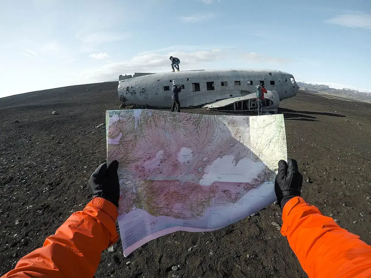 Gloved hands holding open a paper map in front of a plane wreck on black sand.
