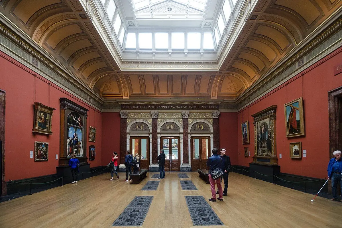 The Central Hall of the National Gallery with old paintings on red walls and a skylight above.