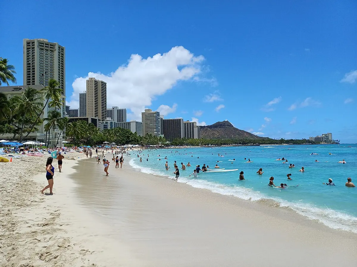 Swimmers in turquoise water along Waikiki Beach, with hotel towers lining the shore and Diamond Head under a blue sky